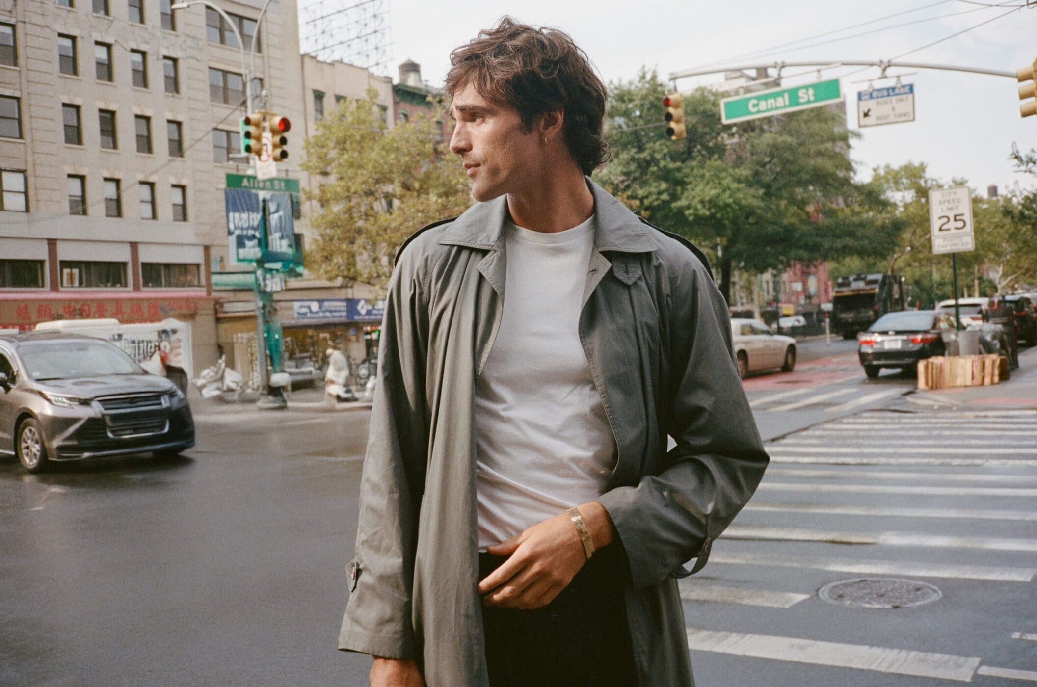 Male model in gray trench coat walks on a crosswalk with city buildings and cars in the background in an urban scene.