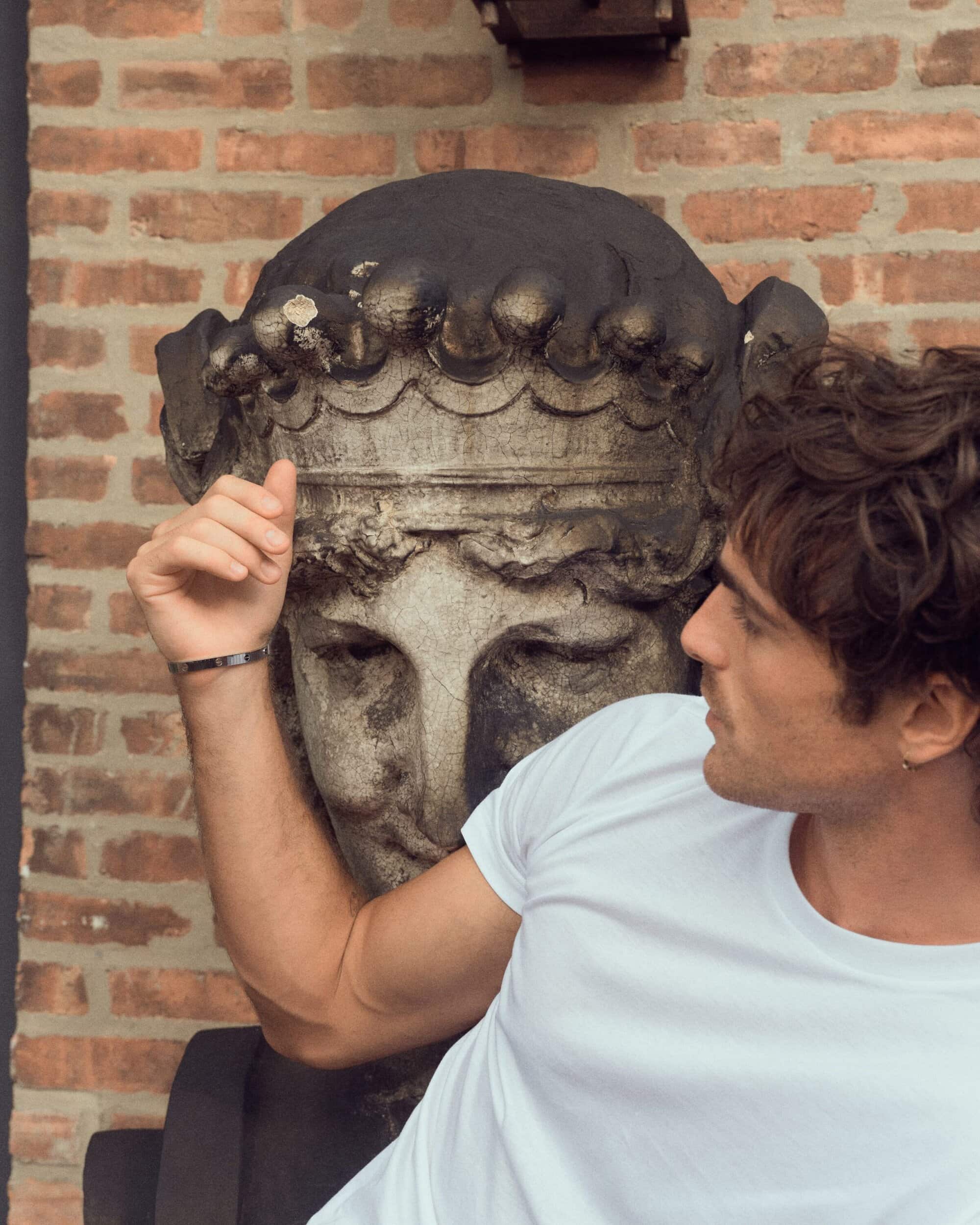 Man in a white t-shirt poses next to a weathered stone bust, with a red brick wall visible in the blurred background.