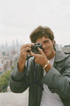 Jacob Elordi wearing a grey jacket and gold cuff bracelet looks through a vintage camera against an urban city skyline background.