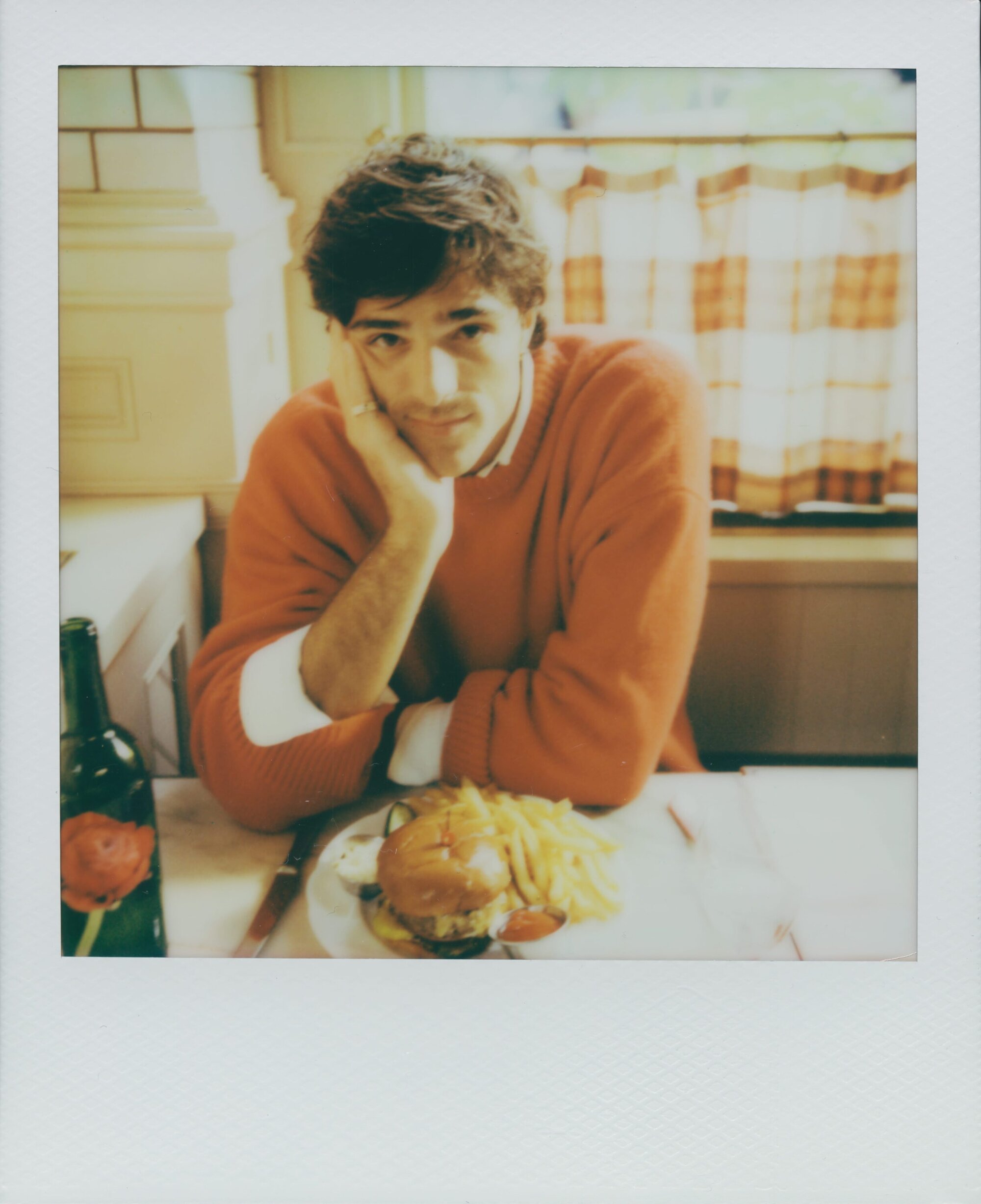 Man in orange sweater at table with burger, fries, and bottle. Casual indoor shot of food and portrait of a young adult male.
