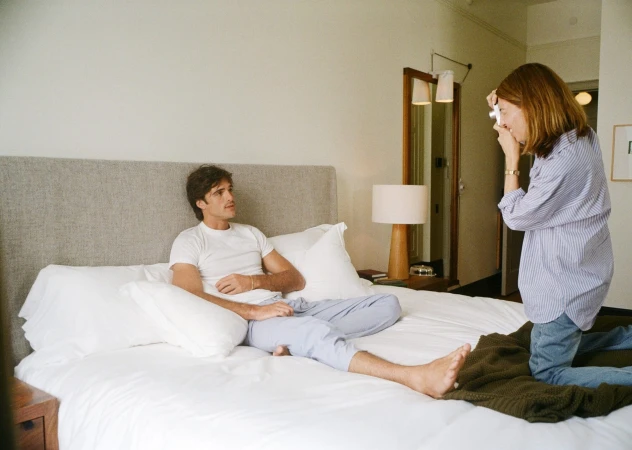 Jacob Elordi in a white t-shirt and light blue trousers sits on a bed while a woman in a striped shirt photographs him with a vintage camera.
