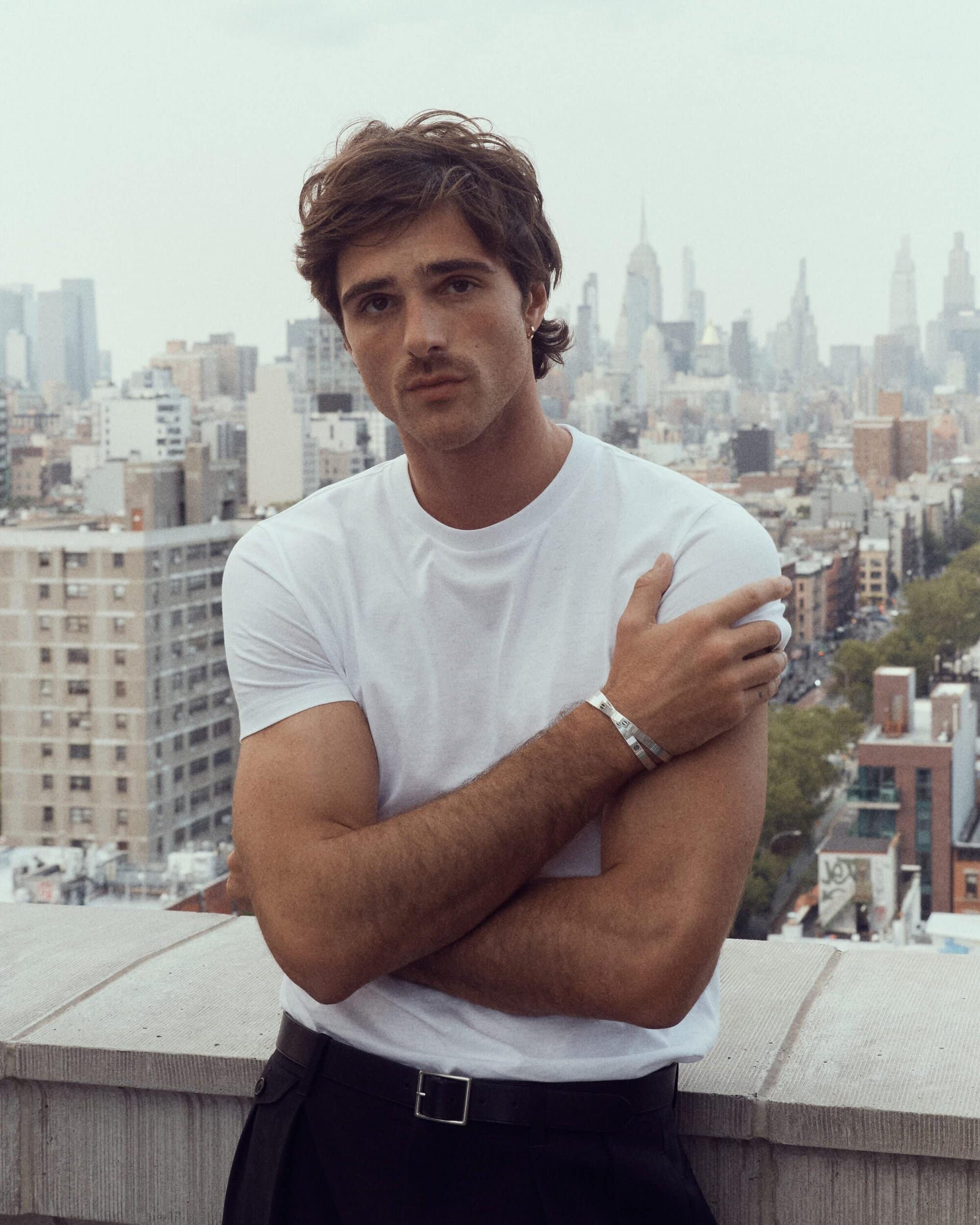 Portrait of a young adult man with crossed arms in front of an urban skyline featuring skyscrapers and an overcast sky.