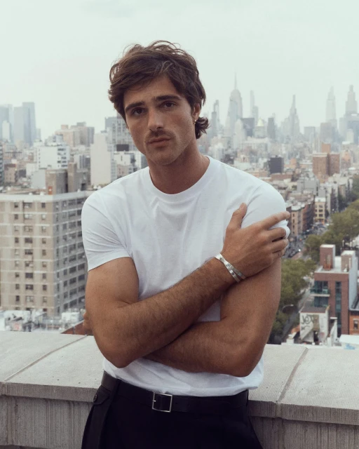 Portrait of a young adult man with crossed arms in front of an urban skyline featuring skyscrapers and an overcast sky.