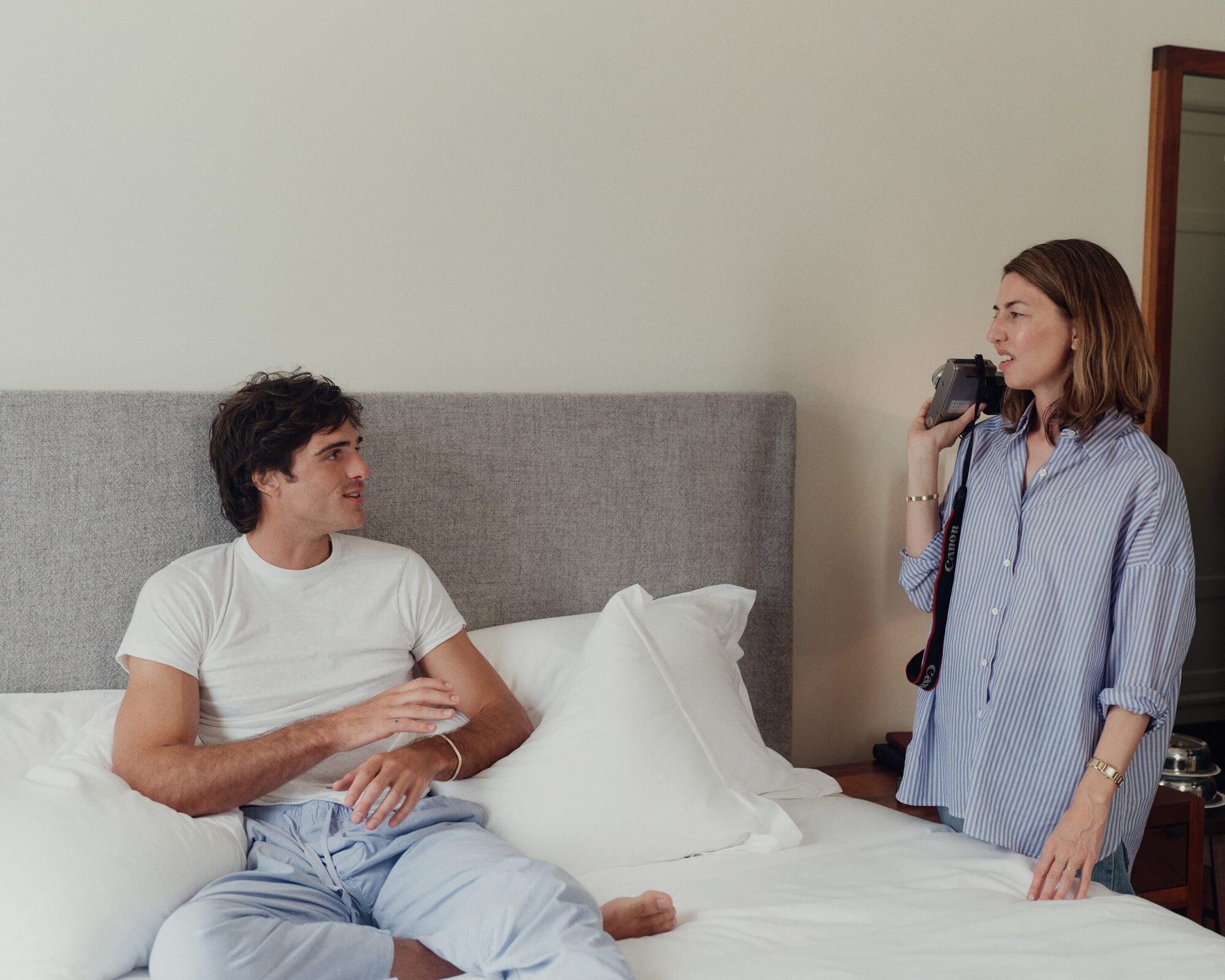 An adult male sits on a bed while an adult female stands nearby holding a camera in an indoor setting with natural lighting.