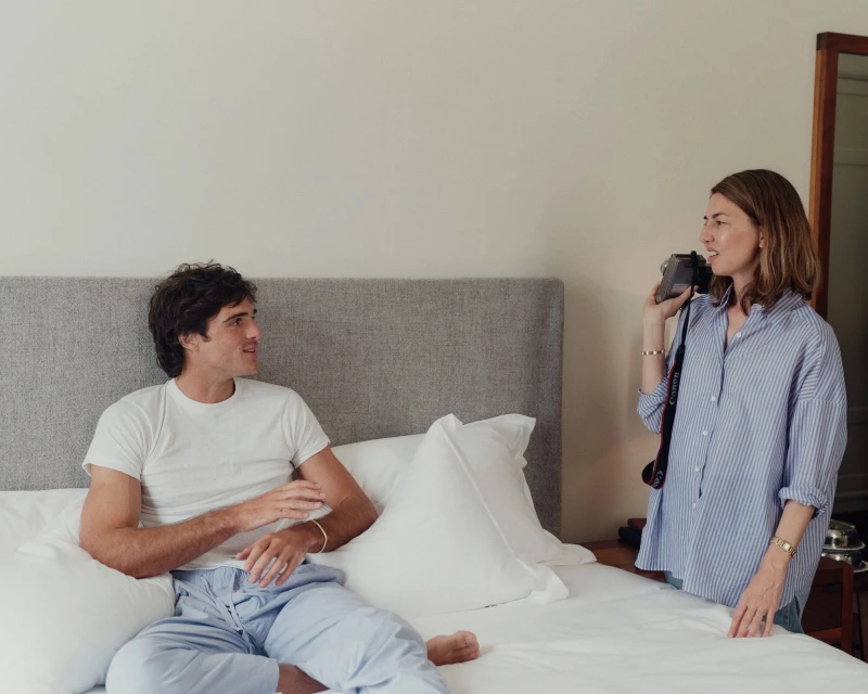An adult male sits on a bed while an adult female stands nearby holding a camera in an indoor setting with natural lighting.