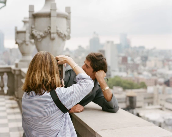 Woman in blue striped shirt holding a vintage camera photographing Jacob Elordi on a stone rooftop overlooking a city.