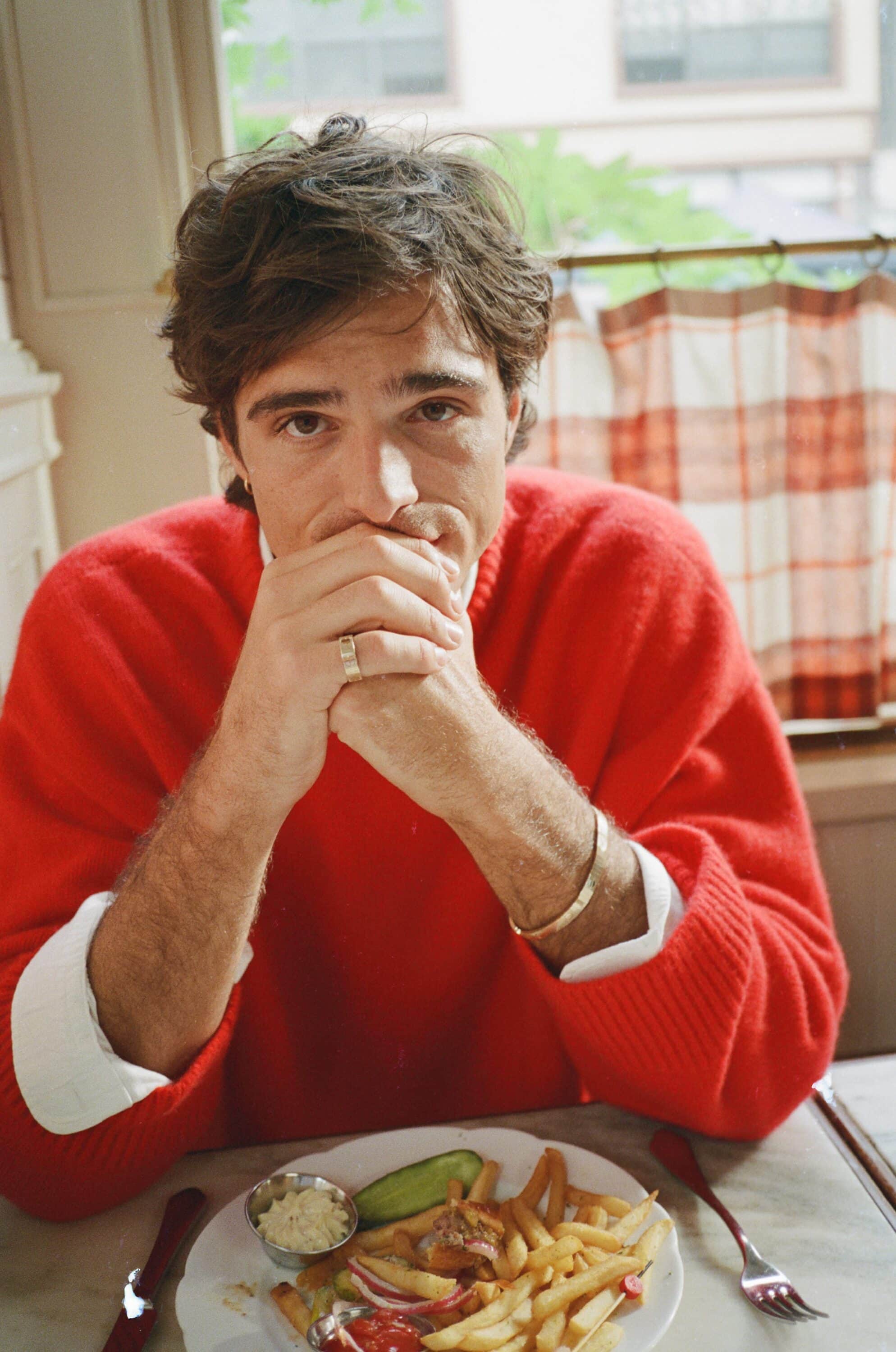Adult male in red sweater sits at a table with food, including fries, in a brightly lit indoor restaurant setting.