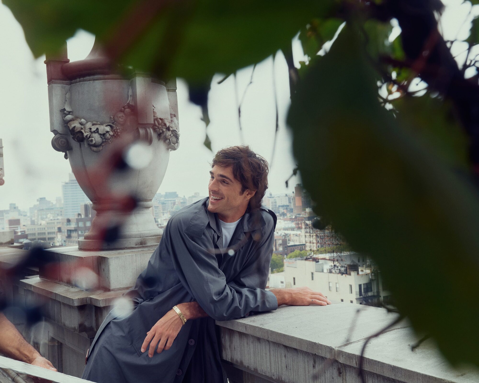 Young man in grey coat smiles on a rooftop with city views, framed by blurry green leaves. Sunny daytime scene.