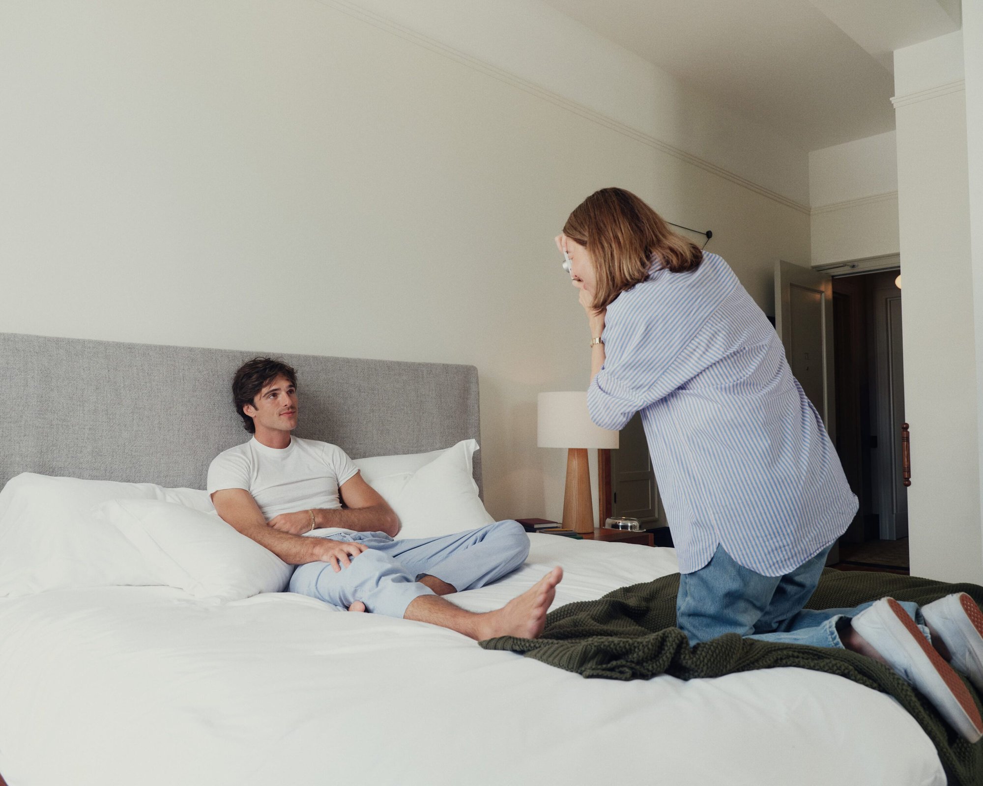 Indoor scene features a man reclining on a bed while a woman kneels at the bedside, creating a candid and relaxed atmosphere.