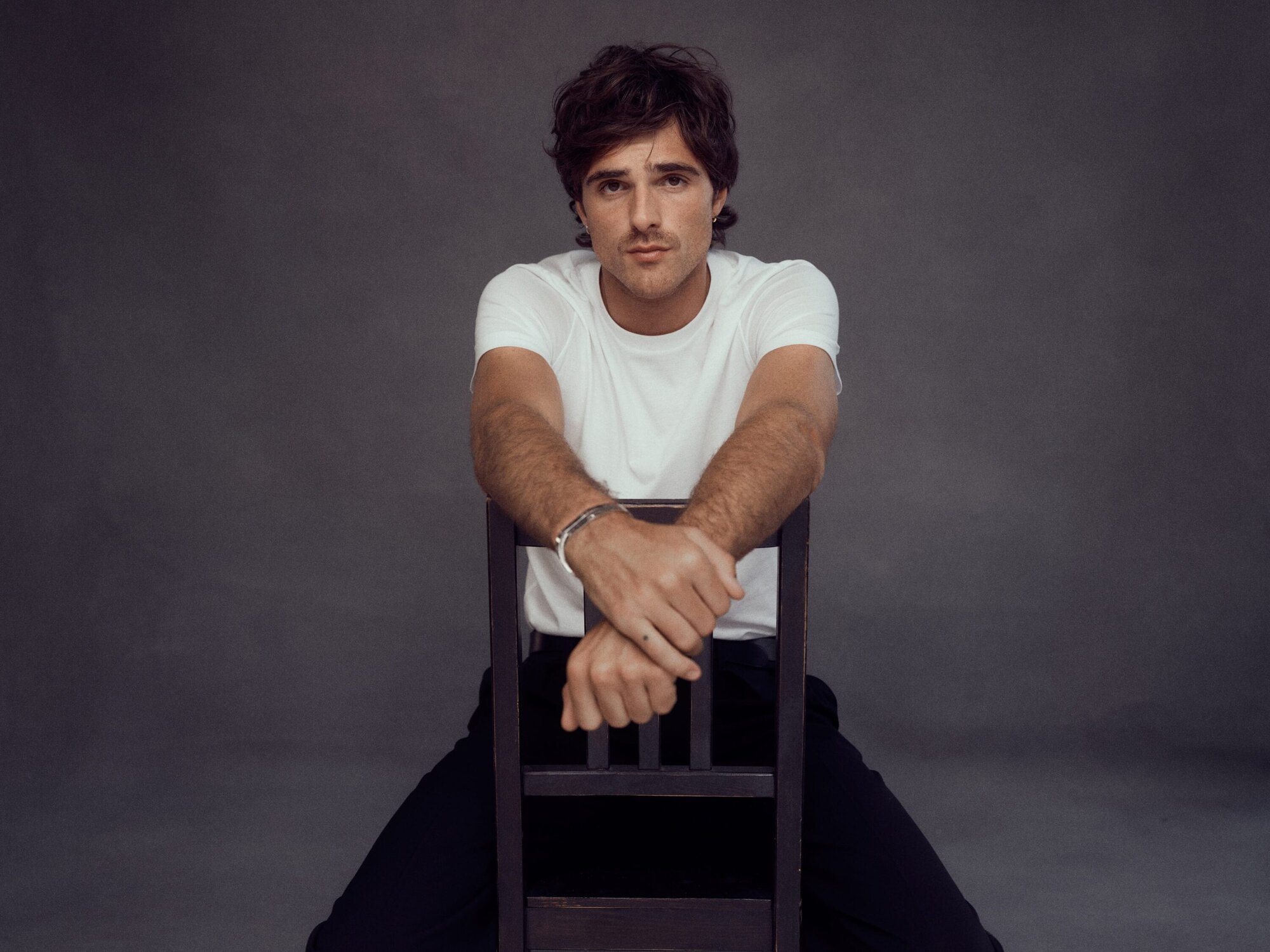 Jacob Elordi wearing a white t-shirt and resting arms on the back of a wooden chair against a dark grey background.