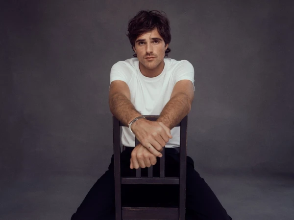 Jacob Elordi wearing a white t-shirt and resting arms on the back of a wooden chair against a dark grey background.