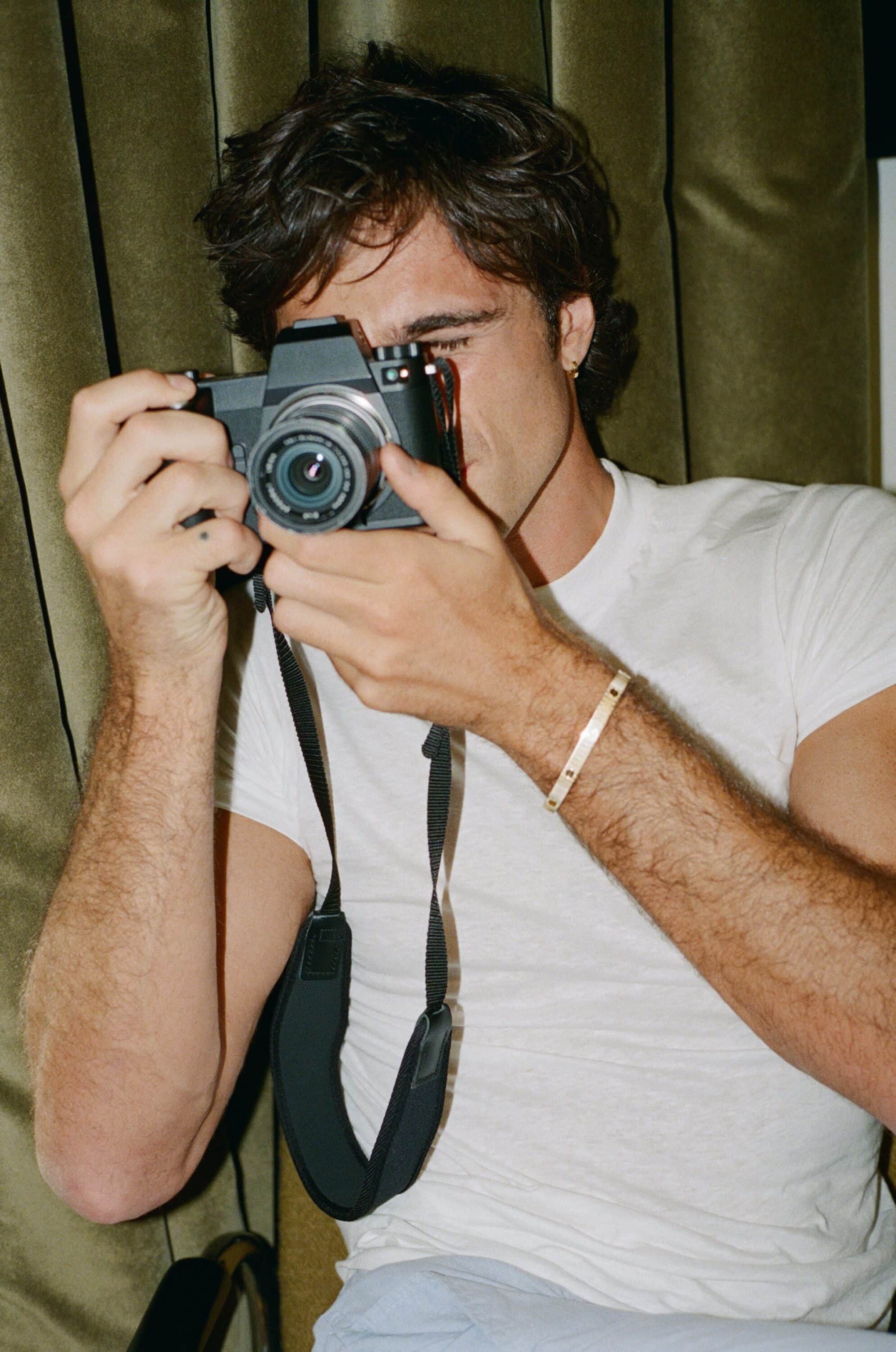 An adult male holds a black digital camera up to his face, standing indoors in front of a fabric backdrop, lit with warm light.