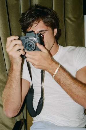 An adult male holds a black digital camera up to his face, standing indoors in front of a fabric backdrop, lit with warm light.