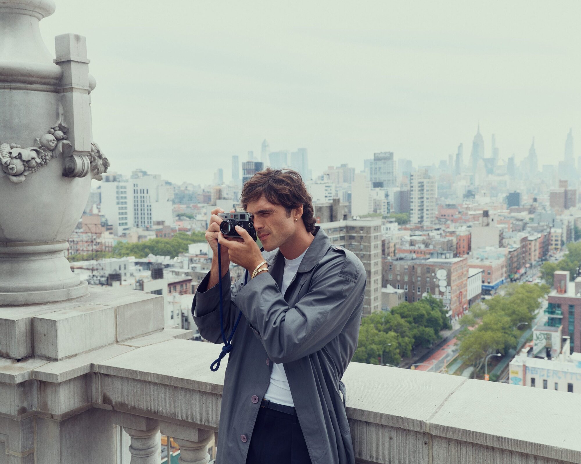 Adult male wearing a trench coat, taking a picture with a film camera on a rooftop with a city skyline view in the background.