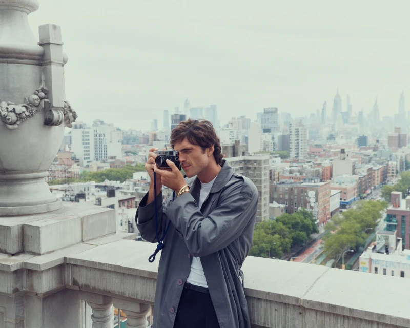 Adult male wearing a trench coat, taking a picture with a film camera on a rooftop with a city skyline view in the background.