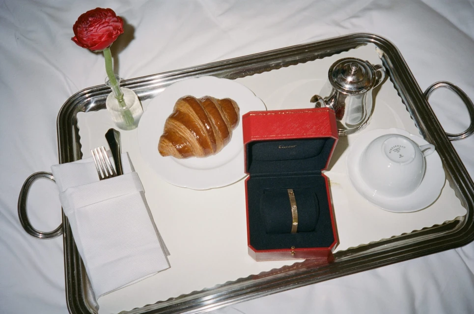 Breakfast in bed featuring a croissant, red flower in a vase, gold bracelet in a red box on a silver tray, and a white bedding backdrop.