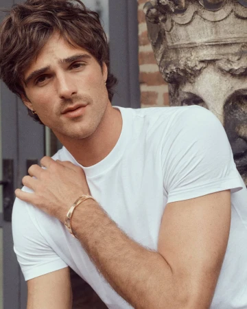 Close-up of handsome adult male wearing a white t-shirt and gold bracelet, posing near an outdoor stone structure in natural light.