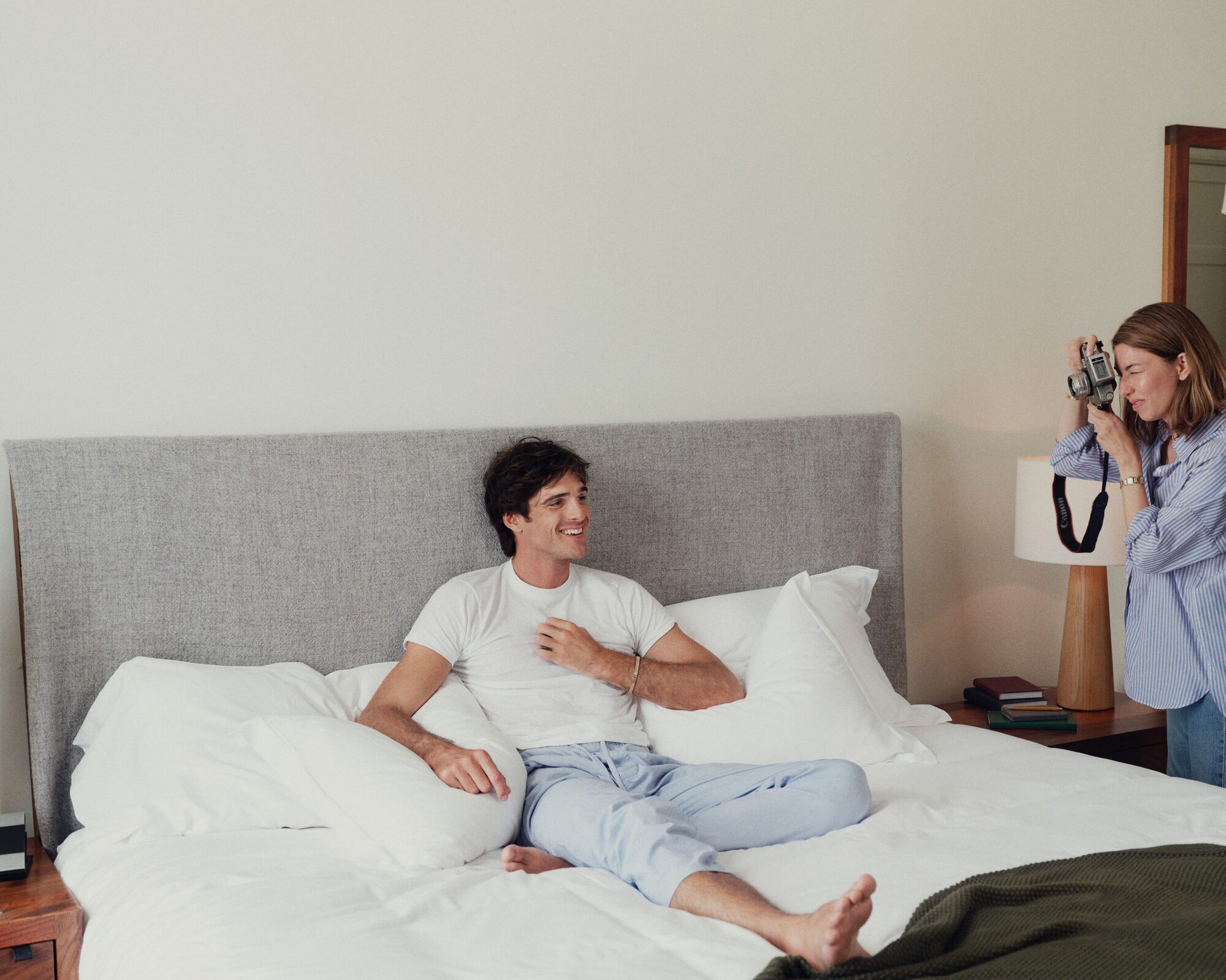 A relaxed man poses on a bed in pajamas as a woman with a camera captures the moment in a bright, casual indoor setting.