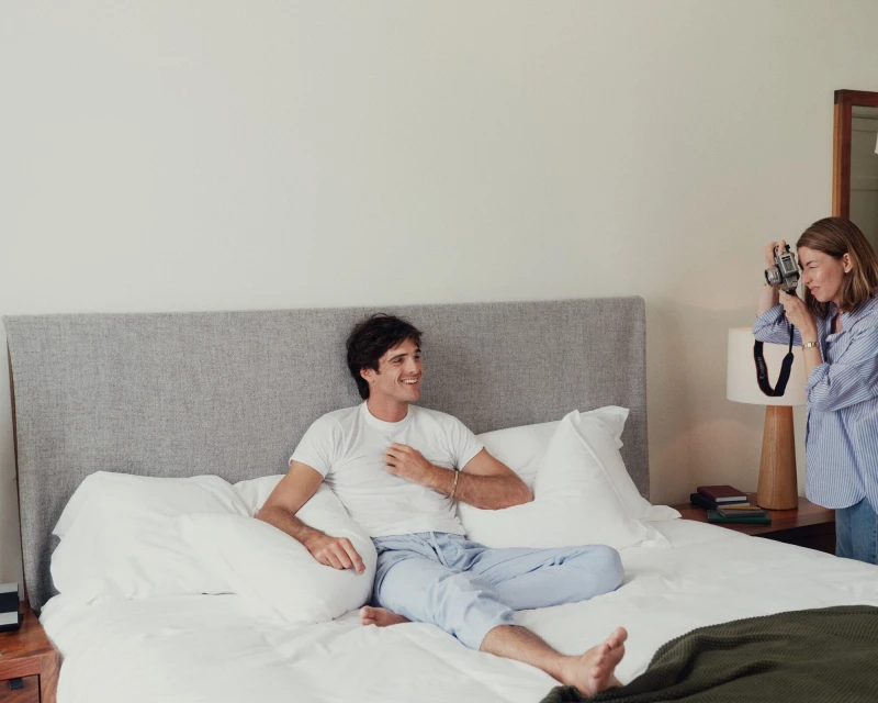 A relaxed man poses on a bed in pajamas as a woman with a camera captures the moment in a bright, casual indoor setting.