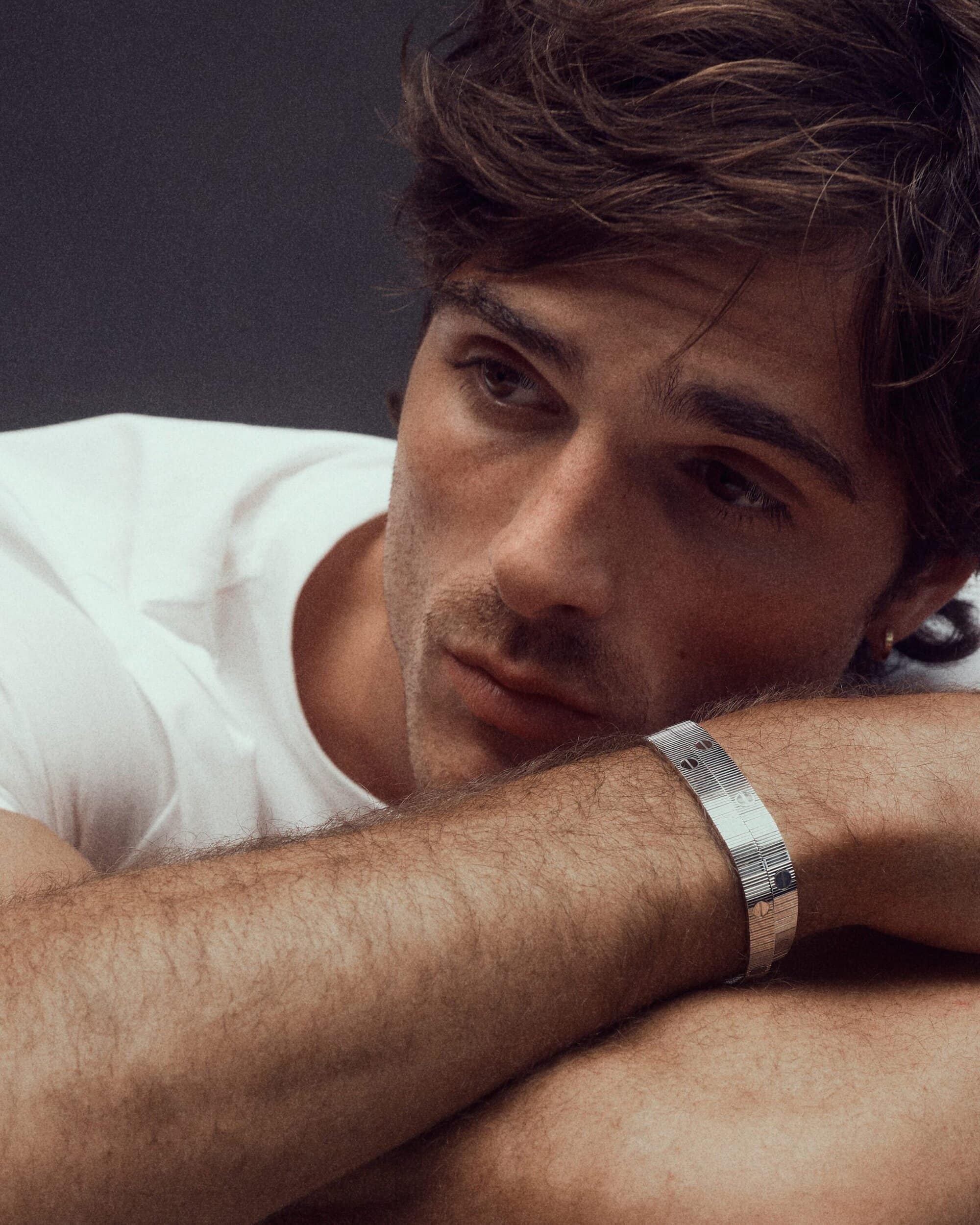 Close-up of a young adult male with brown hair and light skin, resting with his arms crossed, wearing a white t-shirt and silver bracelet.