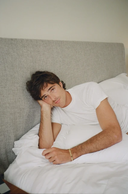 Adult man in a white T-shirt lying on white bed linens and a grey headboard with his head resting on his arm in an indoor setting.