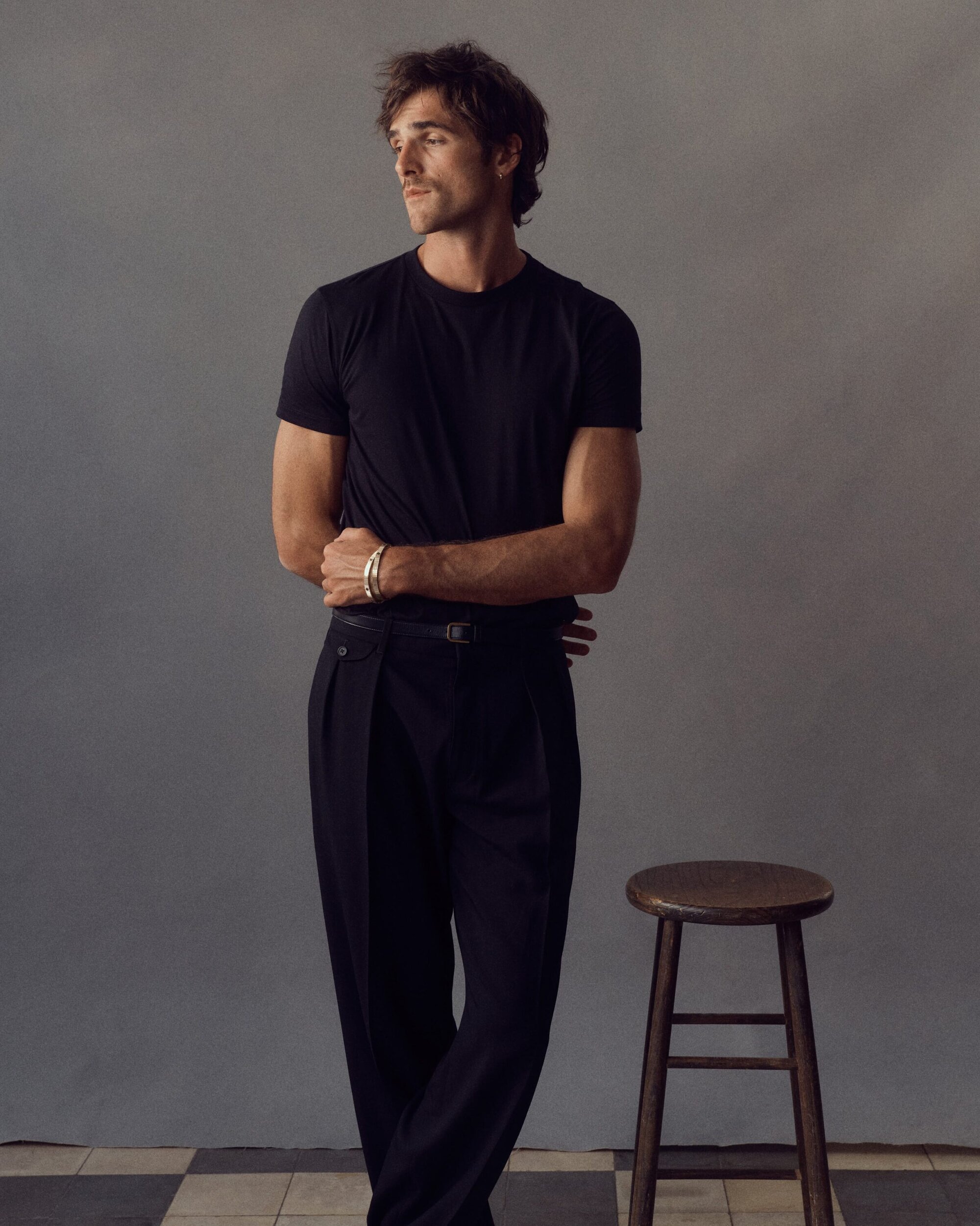Jacob Elordi wearing a black t-shirt and dark trousers standing beside a wooden stool against a grey studio backdrop