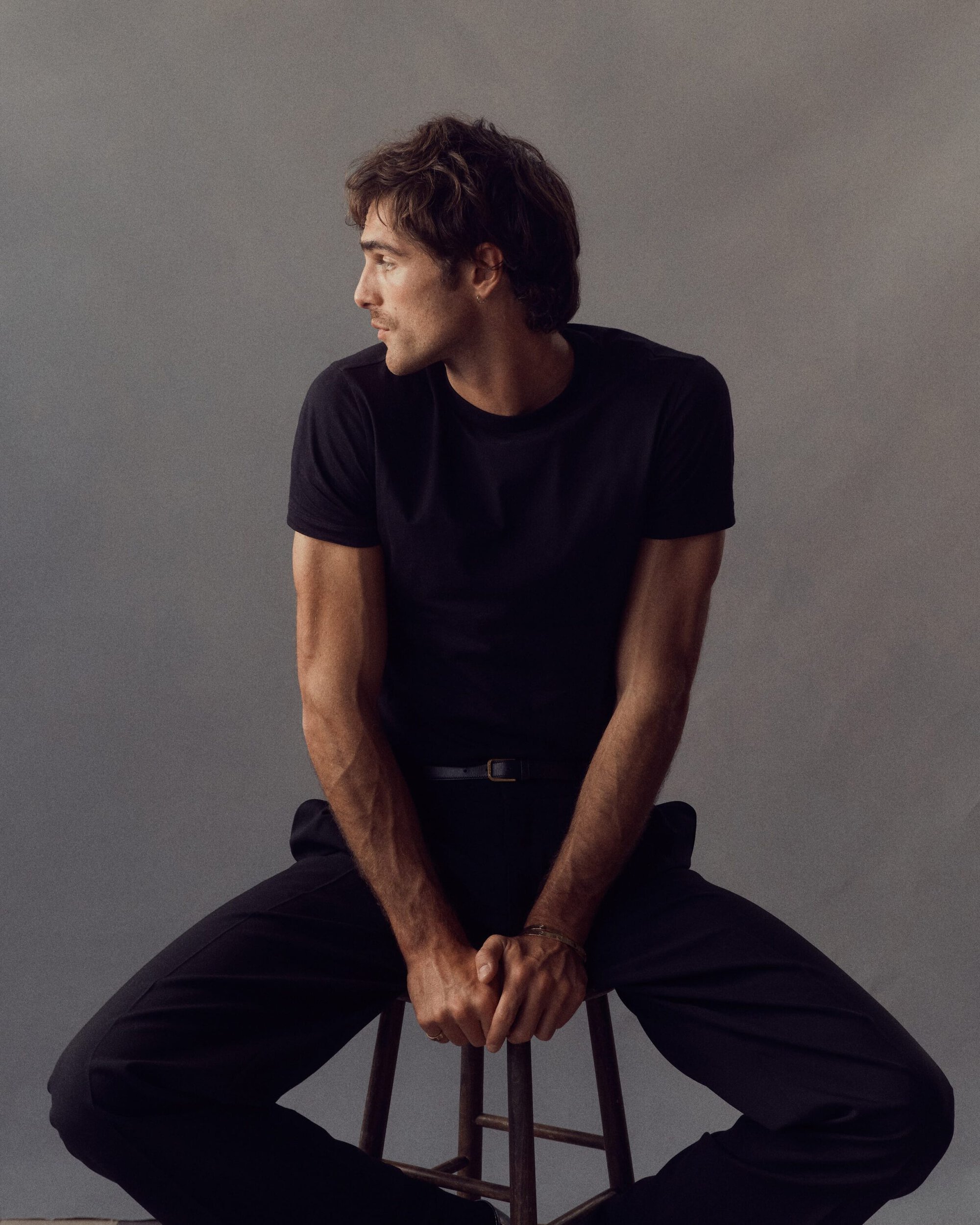 Jacob Elordi poses in a photo studio wearing black pants and a black t-shirt. He is sitting on a wooden stool against a grey background.