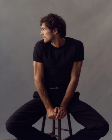 Jacob Elordi poses in a photo studio wearing black pants and a black t-shirt. He is sitting on a wooden stool against a grey background.