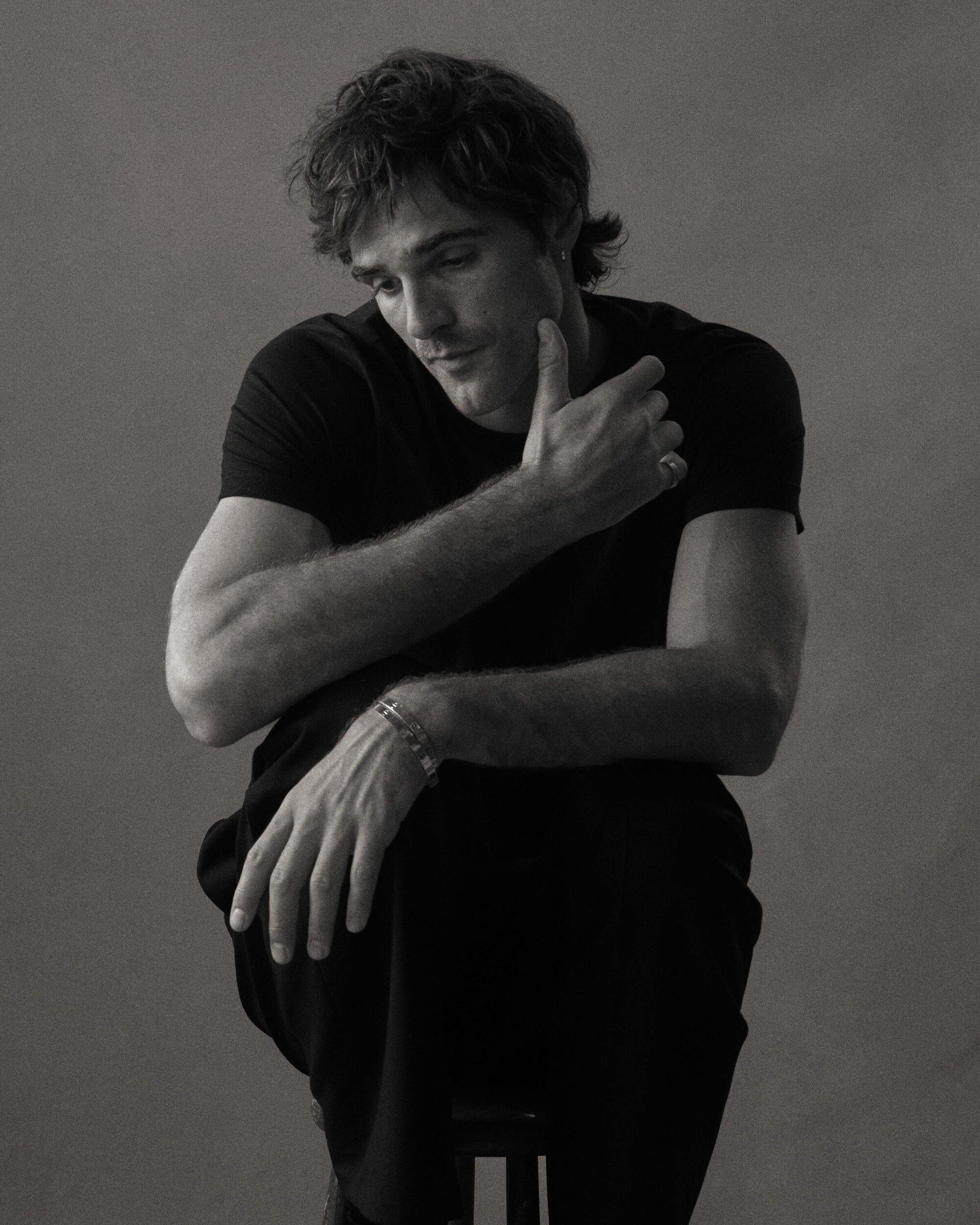 Monochrome portrait of a young adult man sitting on a stool, looking down with his hand to his face in a photo studio.