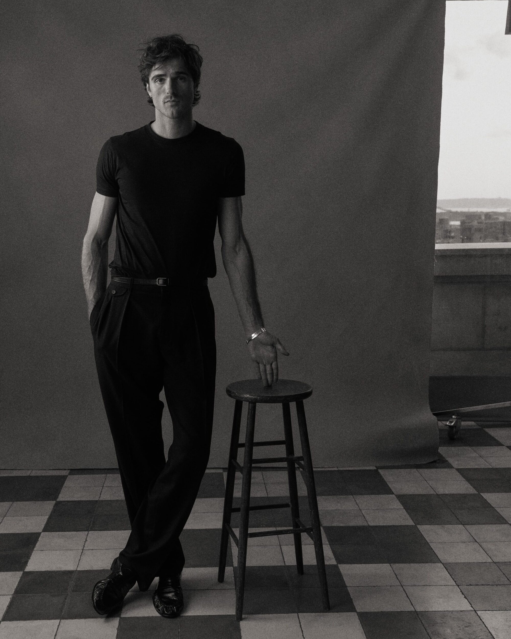Jacob Elordi in a studio portrait, wearing a black t-shirt and trousers, posing with a wooden stool against a gray backdrop.