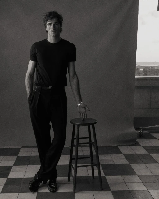 Jacob Elordi in a studio portrait, wearing a black t-shirt and trousers, posing with a wooden stool against a gray backdrop.