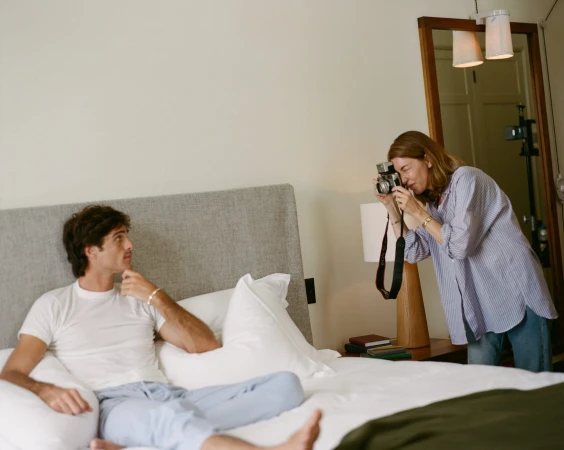 Jacob Elordi seated on a bed in a white t-shirt while photographer Sofia Coppola takes a picture of him with a vintage camera.