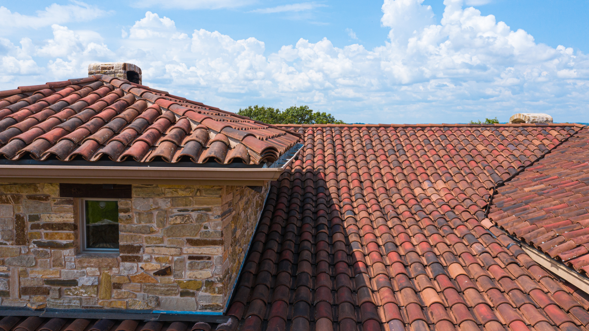 Terracotta tile roof on a stone building under a blue sky with clouds, showcasing architectural design and roofing materials.