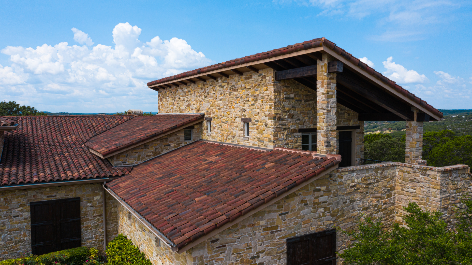 Exterior of a stone building featuring a red tile roof and dark shutters under a partly cloudy sky in a natural landscape.