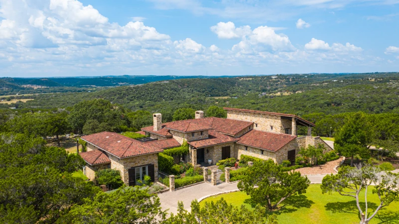 Exterior view of a large tan stone house with a red tile roof, green lawn, and trees, set against a backdrop of rolling hills and a blue sky.