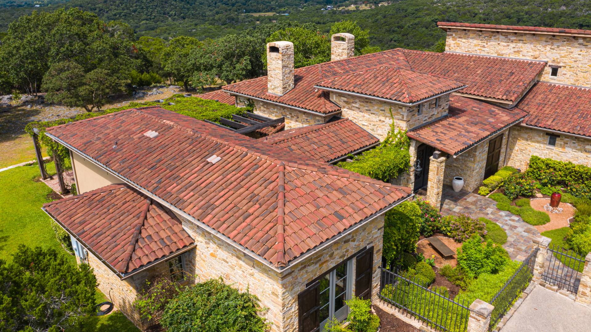 Aerial view of a sandstone house with a terracotta tiled roof, surrounded by lush greenery and trees, on a sunny day.