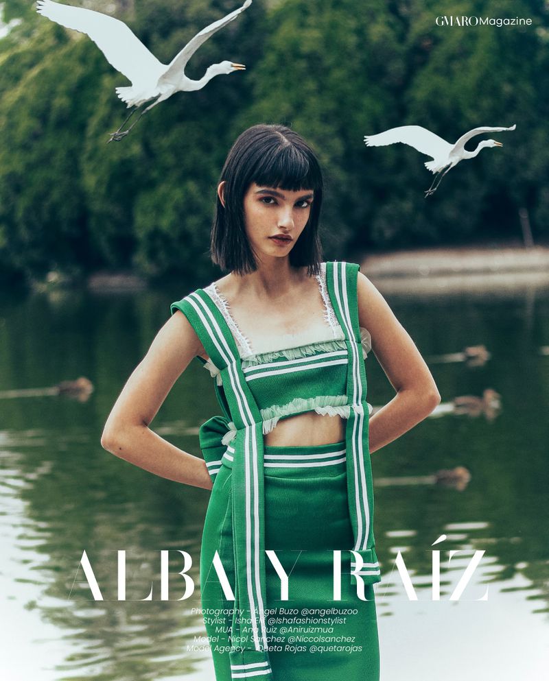 Brunette woman in green outfit posing in front of a lake with white birds flying in the background for a fashion editorial.