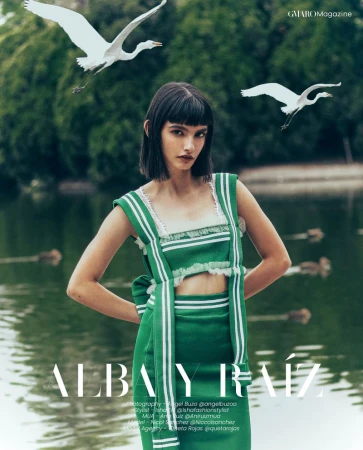 Brunette woman in green outfit posing in front of a lake with white birds flying in the background for a fashion editorial.