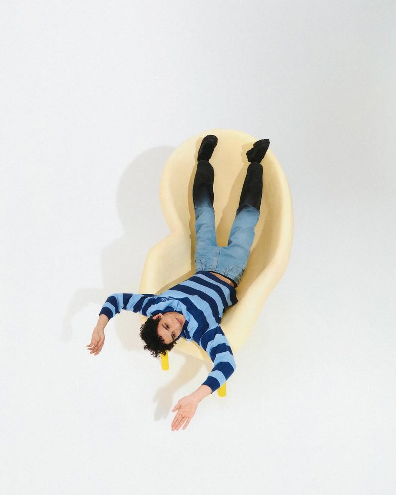 Eye-level studio shot of a man in jeans, striped shirt, and dark socks reclining on a pale yellow chair. Modern, simple composition.