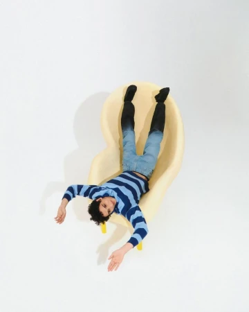 Eye-level studio shot of a man in jeans, striped shirt, and dark socks reclining on a pale yellow chair. Modern, simple composition.