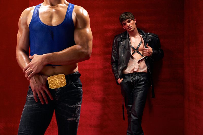 Two adult men pose against a red wall, one wearing a blue tank top and jeans, the other in a leather jacket and pants.