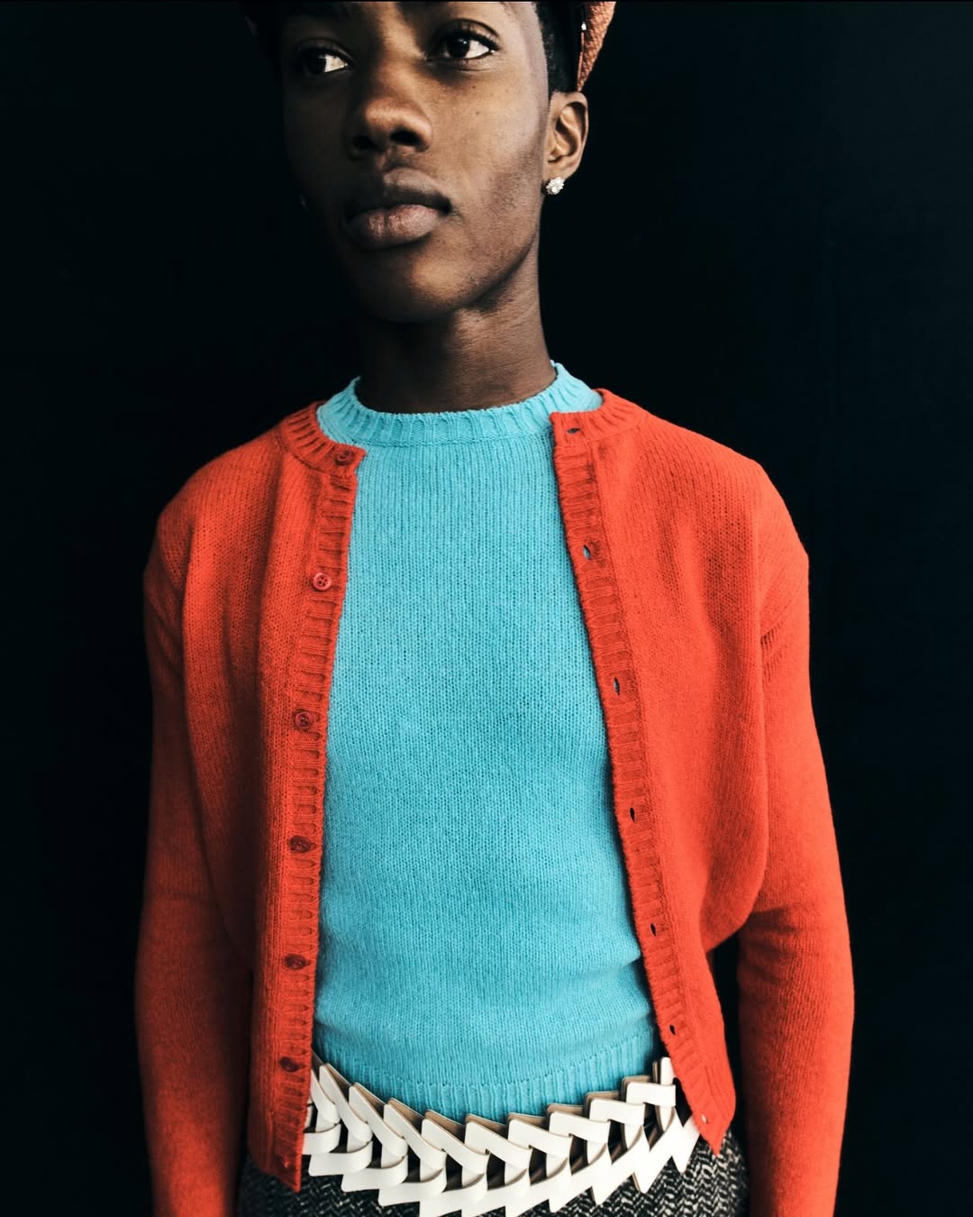Close-up of a black non-binary person wearing a turquoise sweater, orange cardigan and geometric white belt against black background.