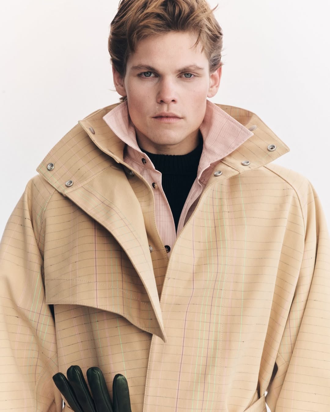 Portrait of a young adult male with blond hair wearing a beige trench coat, gloves, and collared shirt against a white backdrop.
