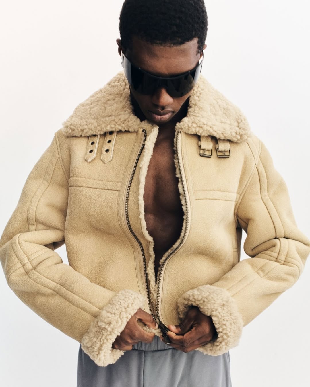 Adult male in a tan shearling bomber jacket with dark glasses, standing against a white background, hands adjusting the zipper.