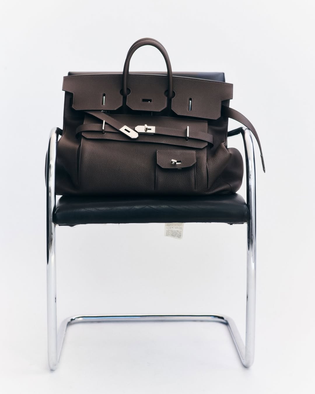 Luxury brown leather Birkin handbag by Hermès sitting on a black leather and chrome chair against a white background in a product shot.