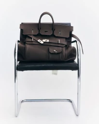 Brown leather handbag resting on a black and chrome modern chair against a bright white background.