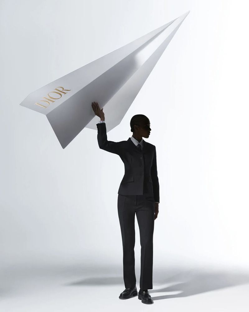 A person in a dark business suit stands in a photo studio holding a large paper airplane with the word DIOR printed on the side.