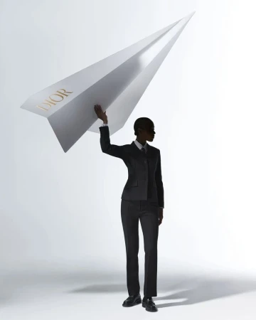 A person in a dark business suit stands in a photo studio holding a large paper airplane with the word DIOR printed on the side.
