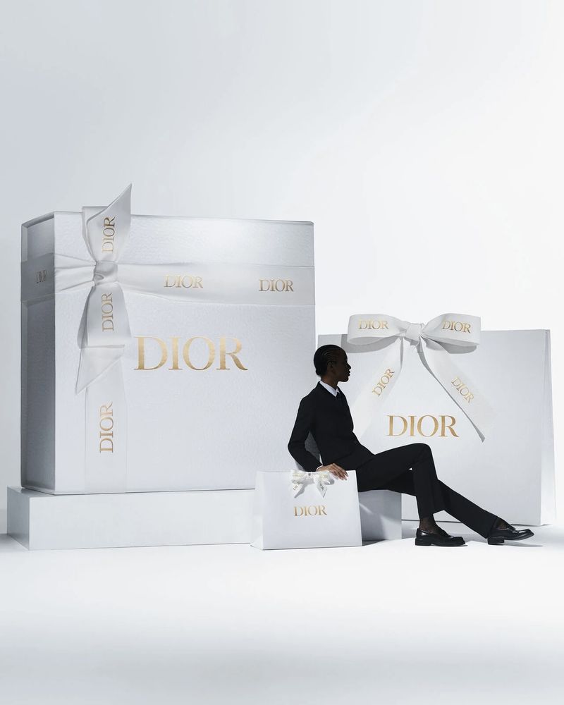 An adult male is sitting next to Dior-branded gift boxes and a shopping bag against a white backdrop, conveying luxury and sophistication.
