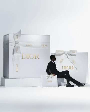 An adult male is sitting next to Dior-branded gift boxes and a shopping bag against a white backdrop, conveying luxury and sophistication.